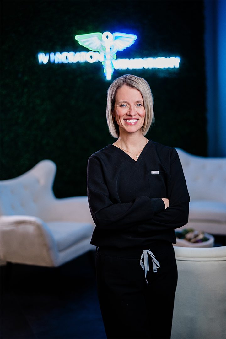 A smiling woman in professional attire standing in front of a neon sign for an iv infusion therapy clinic.